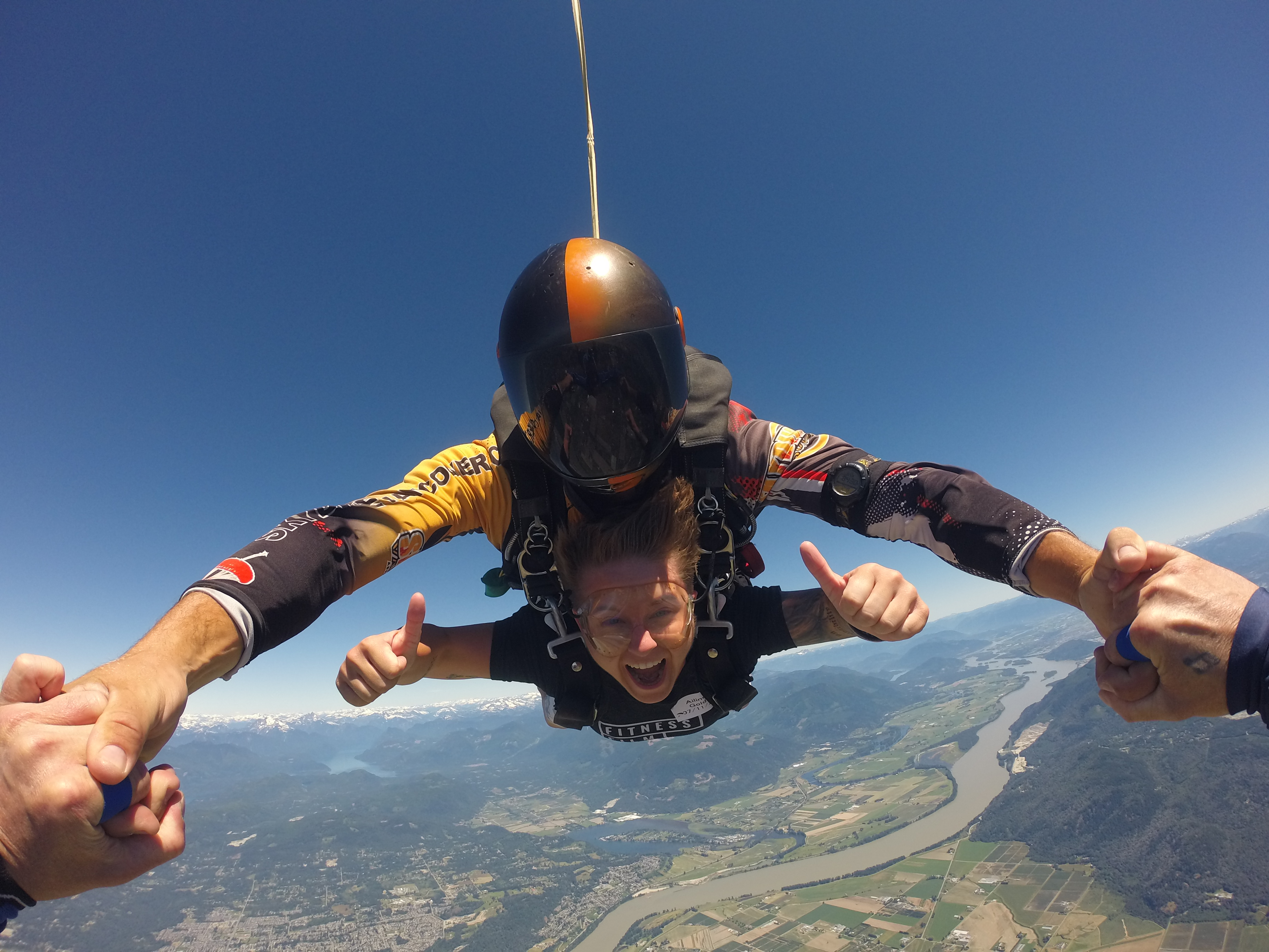 Een jonge vrouw is aan het parachutespringen. Ze glimlacht in de camera en toont duimen omhoog