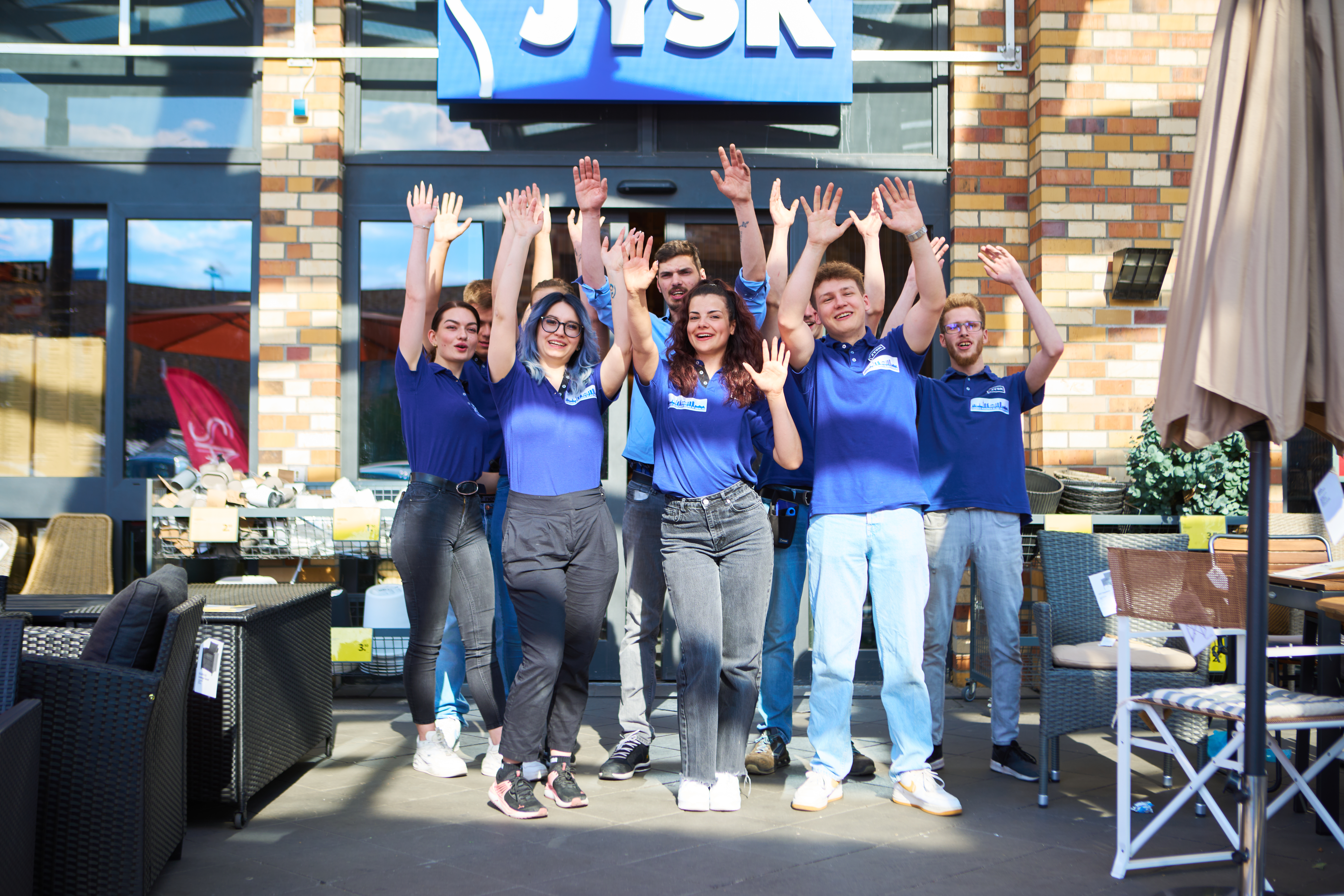 Young people are standing in front of a JYSK store and are lifting theirs arms in the air 
