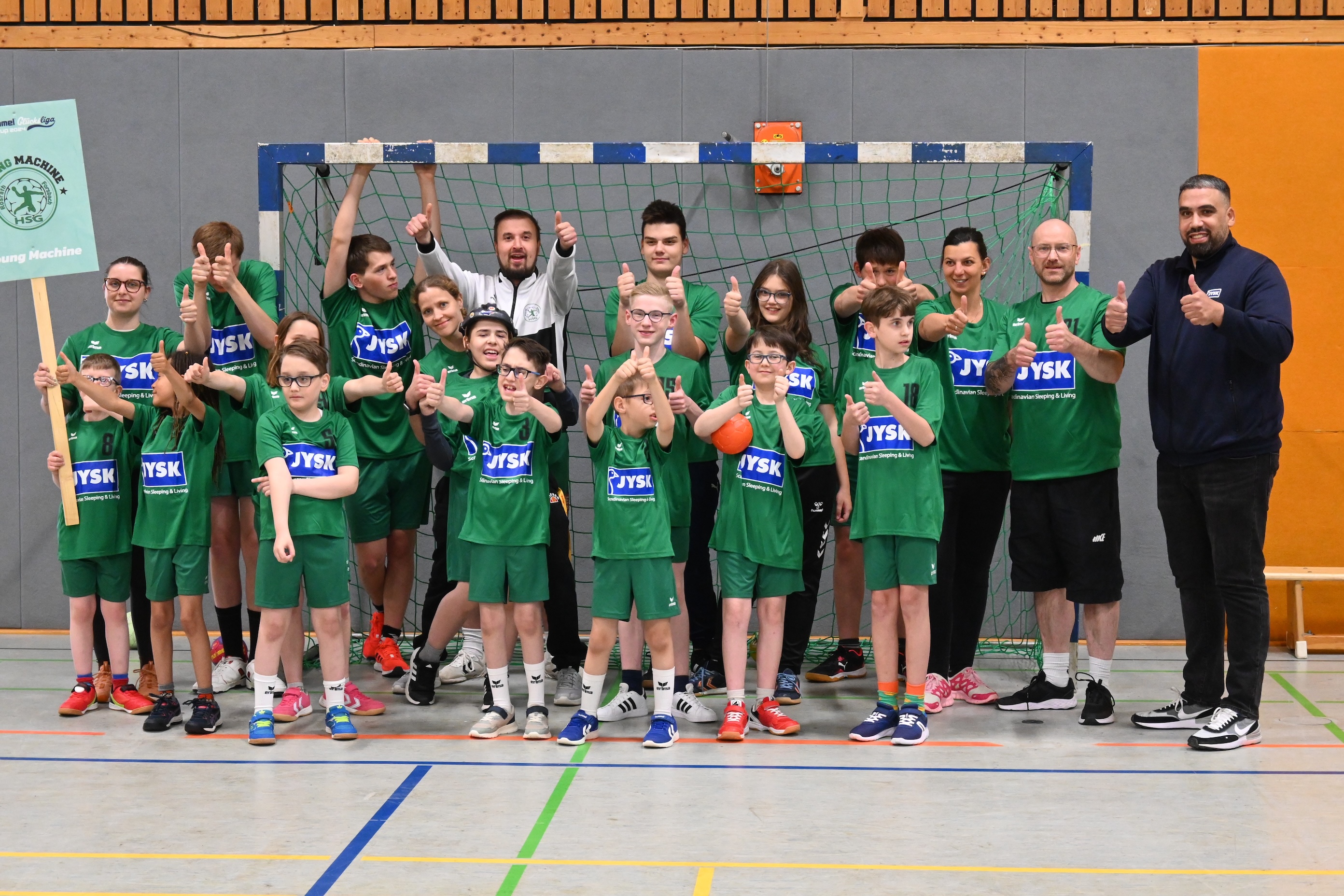 Viele Kinder und zwei Erwachsene stehen in einem Handballtor und jubeln in die Kamera