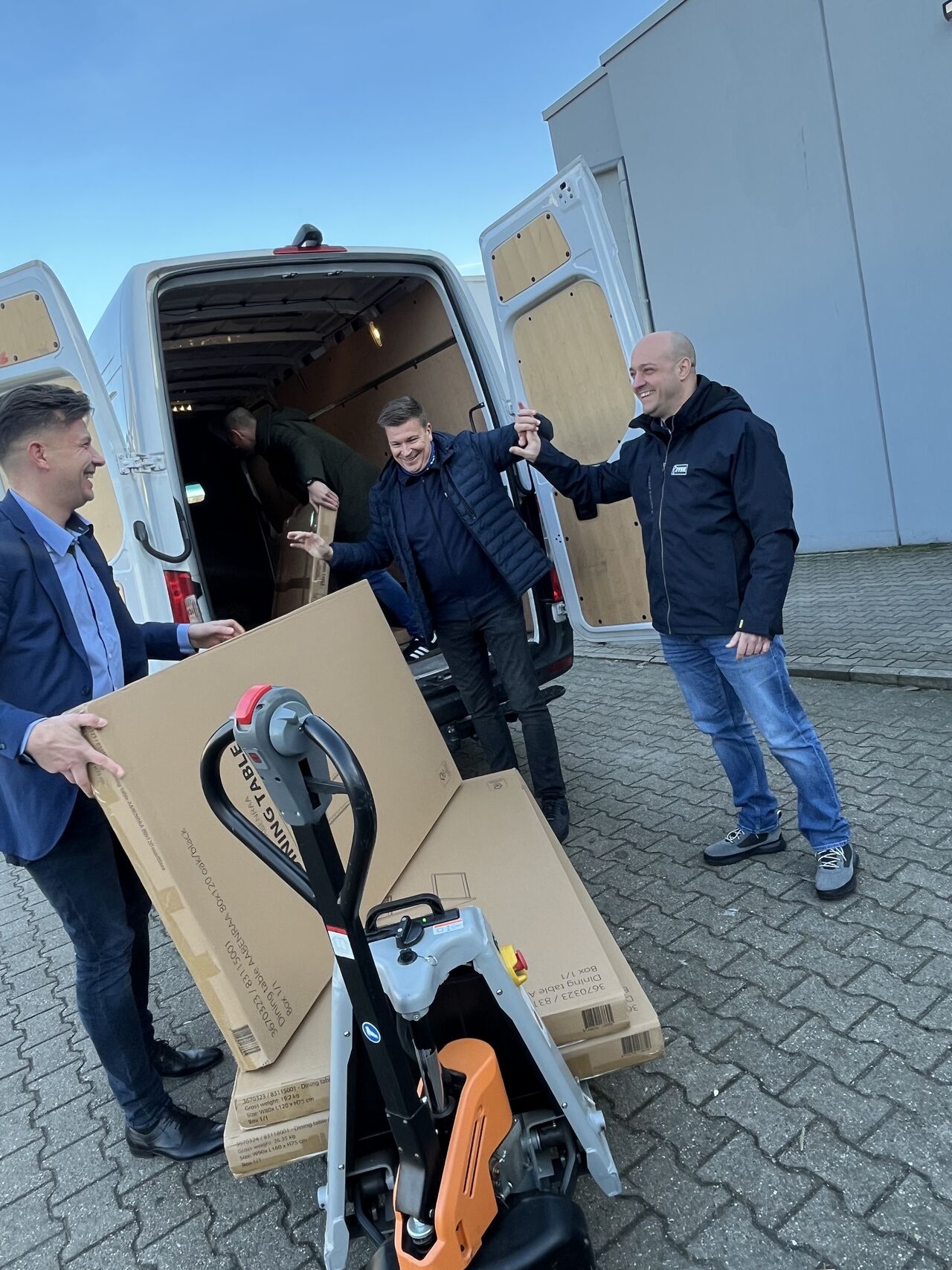 Drei Männer stehen vor einem Kleintransporter und lachen. Der eine Mann hält einen großen Karton fest.
