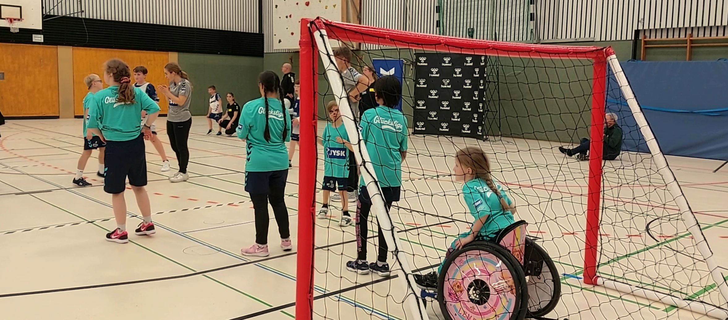 Mehrere Kinder stehen auf einem Handballfeld, im Tor sitzt ein kleines Mädchen mit Pferdeschwanz in einem Rollstuhl und verfolgt konzentriert das Spiel 