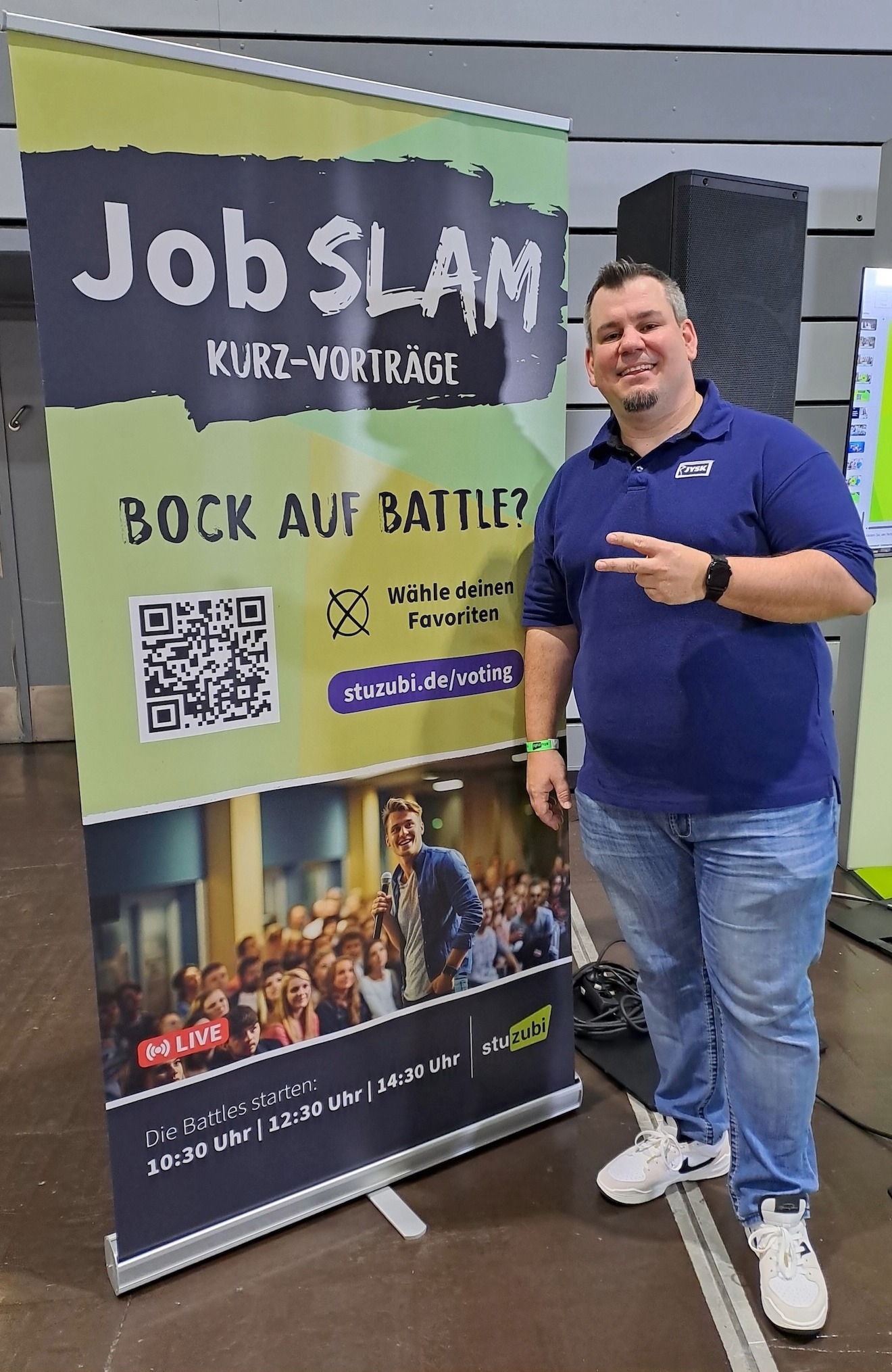 Ein Mann in JYSK-Poloshirt steht vor einem Rollup und lächelt in die Kamera