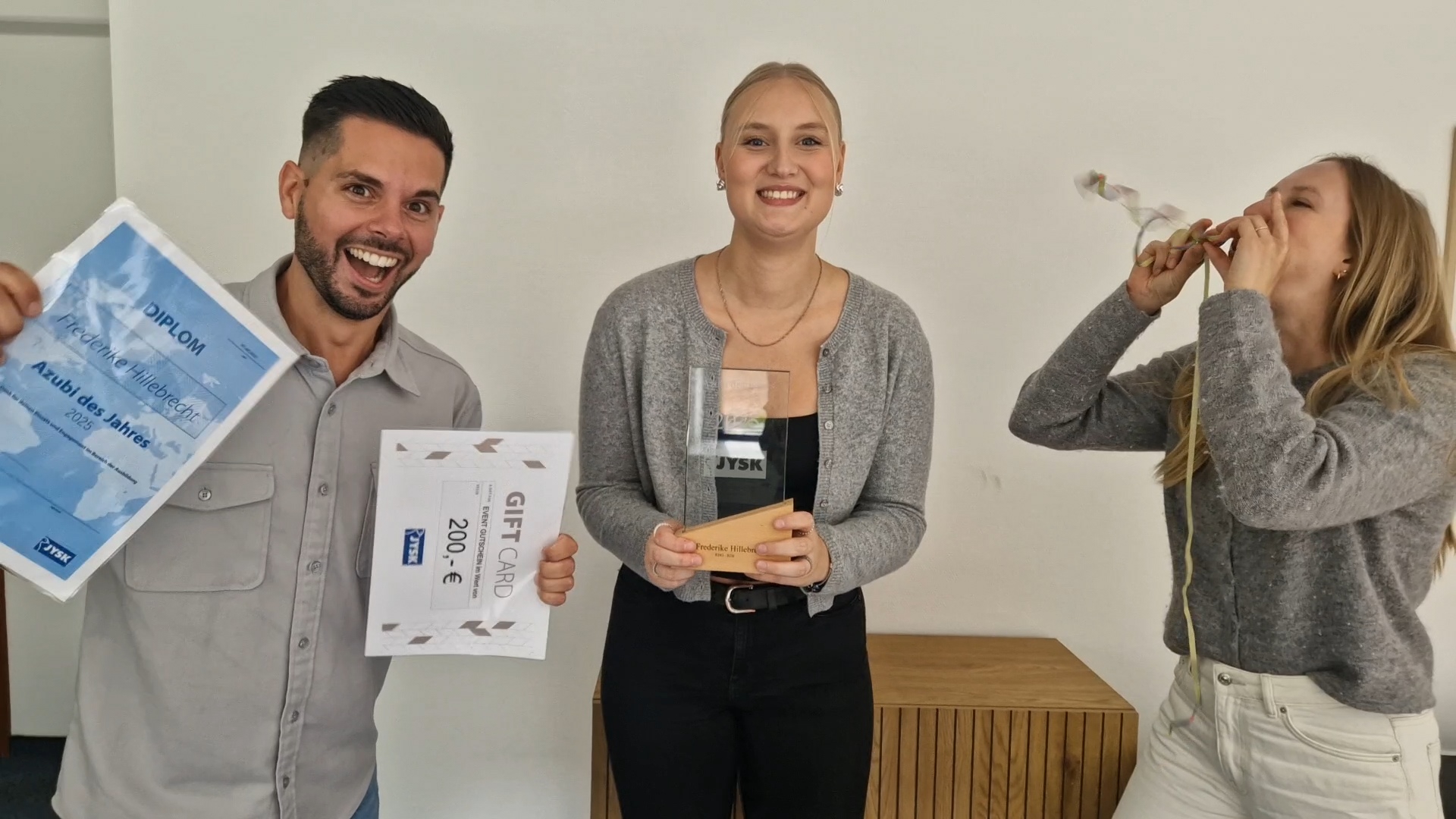 Ein Mann und zwei Frau lachen in die Kamera. Die Frau in der Mitte hält einen Pokal in der Hand. Die andere Frau pustet eine Luftschlange in ihre Richtung. Der Mann hält eine Urkunde und eine Geschenkkarte in den Händen.