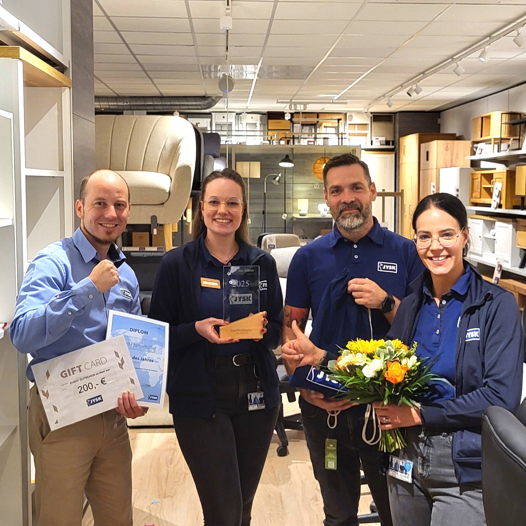 Zwei Männer und zwei Frauen stehen in einem JYSK Store. Ein Mann hält eine urkunde und einen Gutschein in der hand. Eine Frau hält einen Pokal in den Händen. Die zweite Frau hält einen Blumenstrauß in den Händen. .