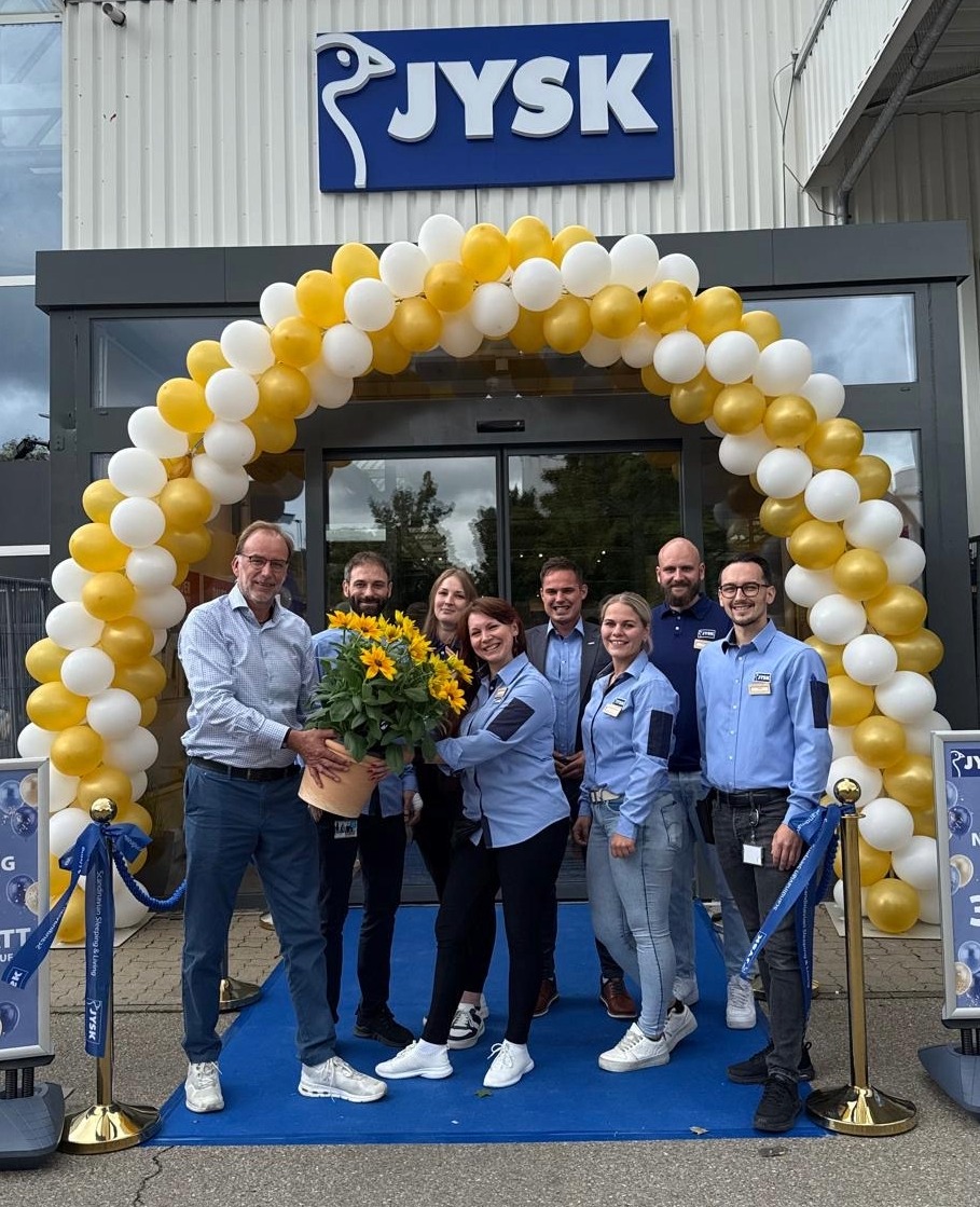 Acht Menschen stehen vor einem JYSK Store. Hinter ihnen ist ein Luftballonbogen zu sehen. Ein Mann reicht einer Frau Blumen