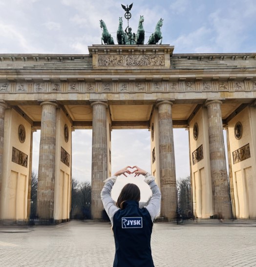 Eine junge Frau mit langen Haaren steht mit dem Rücken zur Kamera, vor ihr erstreckt sich das Brandenburger Tor. mit den Händen formt sie ein Herz. Auf der Weste, welche sie trägt, steht das JYSK-Logo