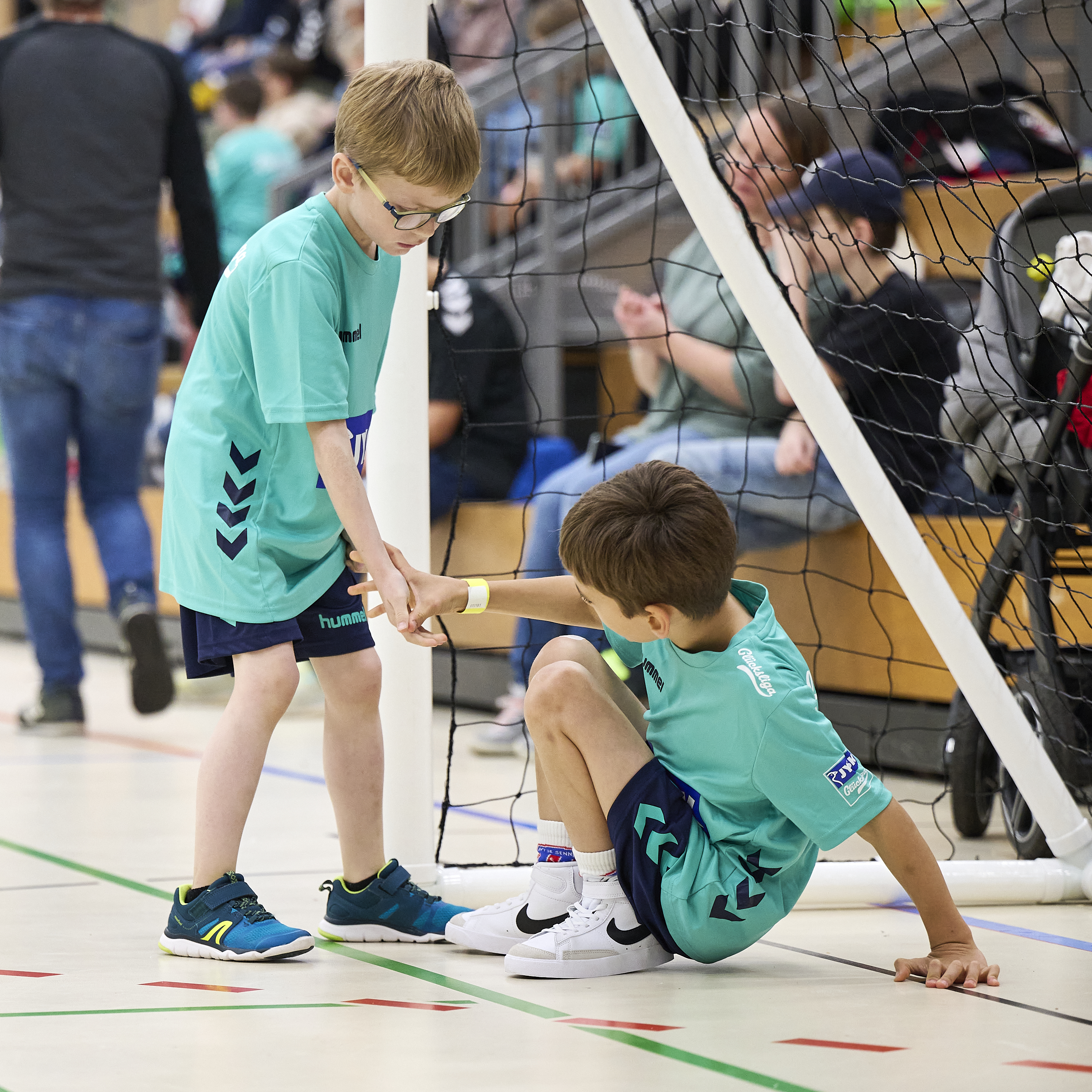 Ein Junge steht vor einem Handballtor. Er reicht einem weiteren Jungen, der auf dem Boden sitzt die Hand und will ihm hochhelfen. 