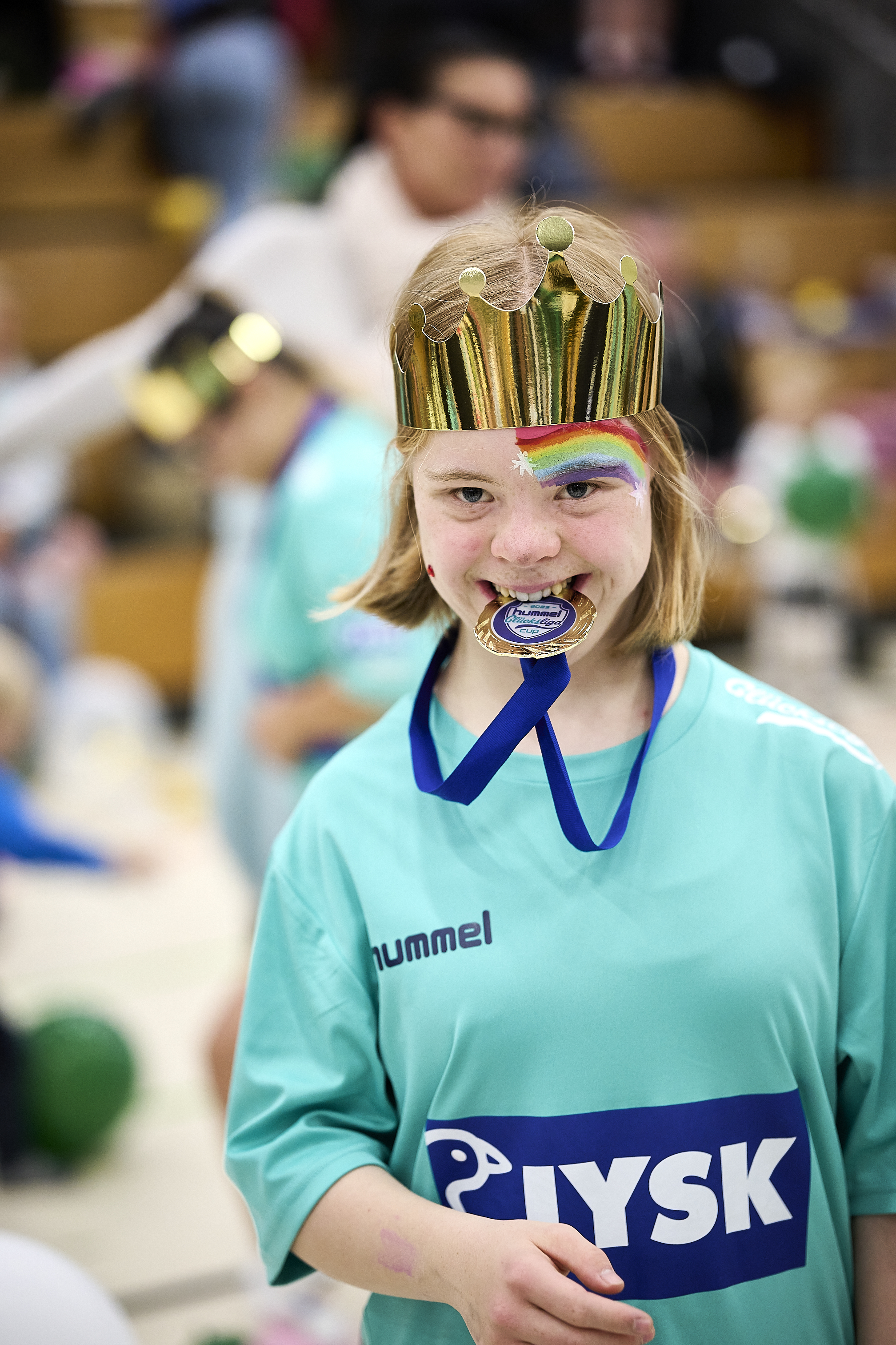 Ein kleines Mädchen trägt eine Papierkrone auf dem Kopf. Sie beißt auf eine Medaille. Sie trägt ein Glücksliga-Trikot und in ihrem Gesicht befindet sich ein geschminkter Regenbogen 