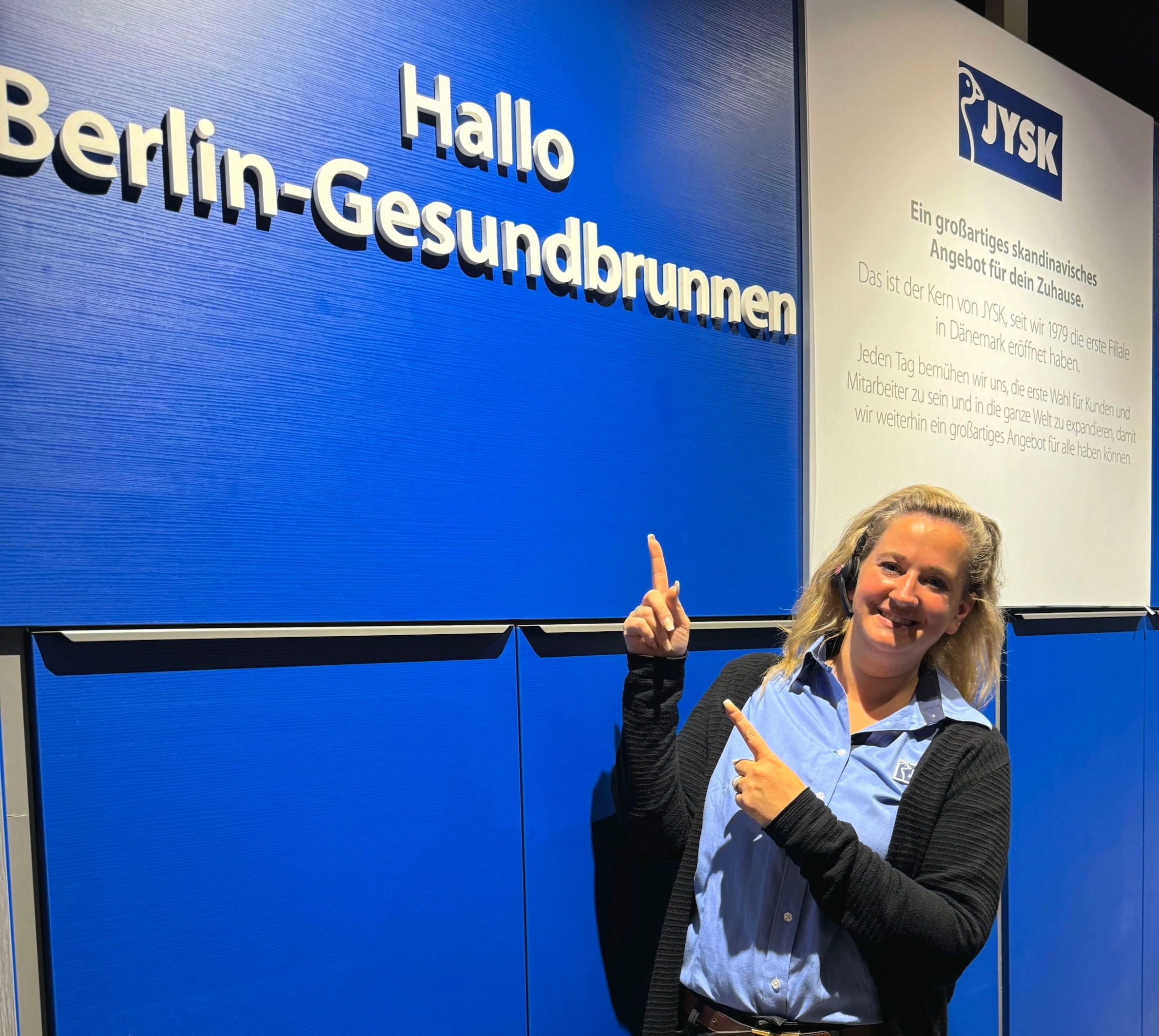 Eine Frau mit hellen, halblangen Haaren trägt JYSK-Arbeitskleidung. Sie zeigt auf den Schriftzug "Hallo Berlin-Gesundbrunnen"