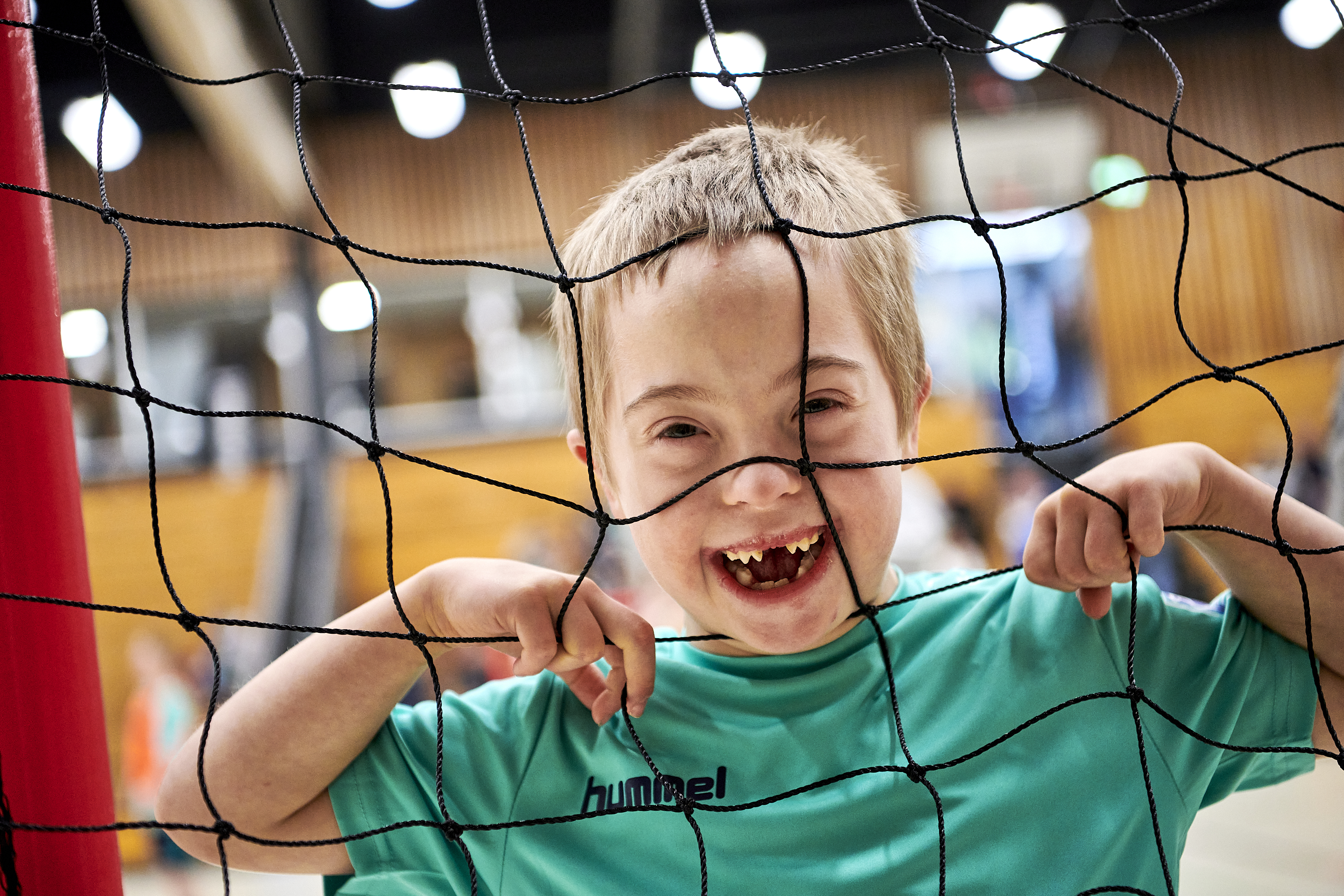 Ein Junge mit kurzen Haaren steht in einem Handballtor und schaut durch das Netz in die Kamera. Er lacht ganz doll und zeigt eine große Zahnlücke 