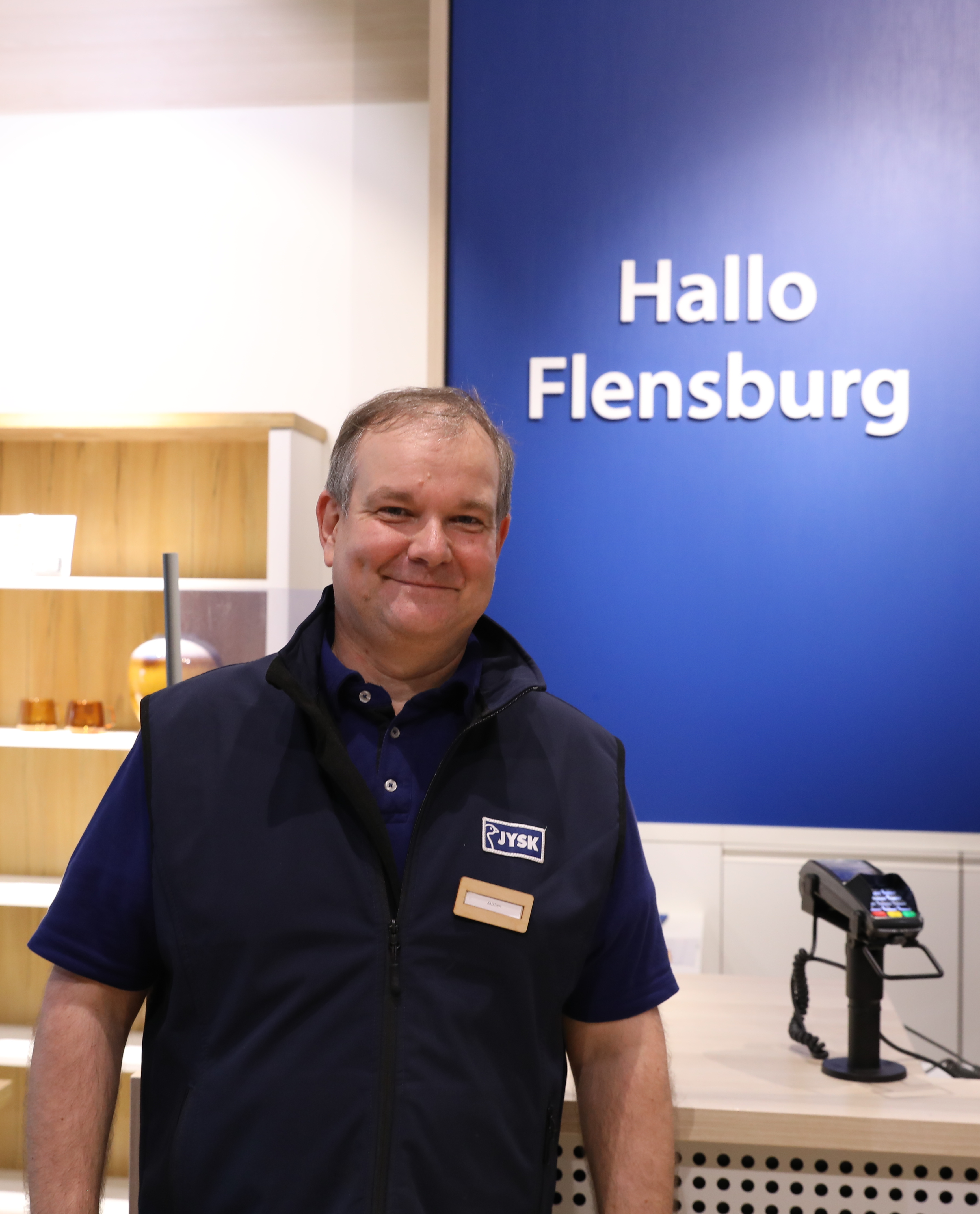 Ein Mann steht vor einem Kassentresen. Im Hintergrund sind die Worte "Hallo Flensburg" zu lesen 