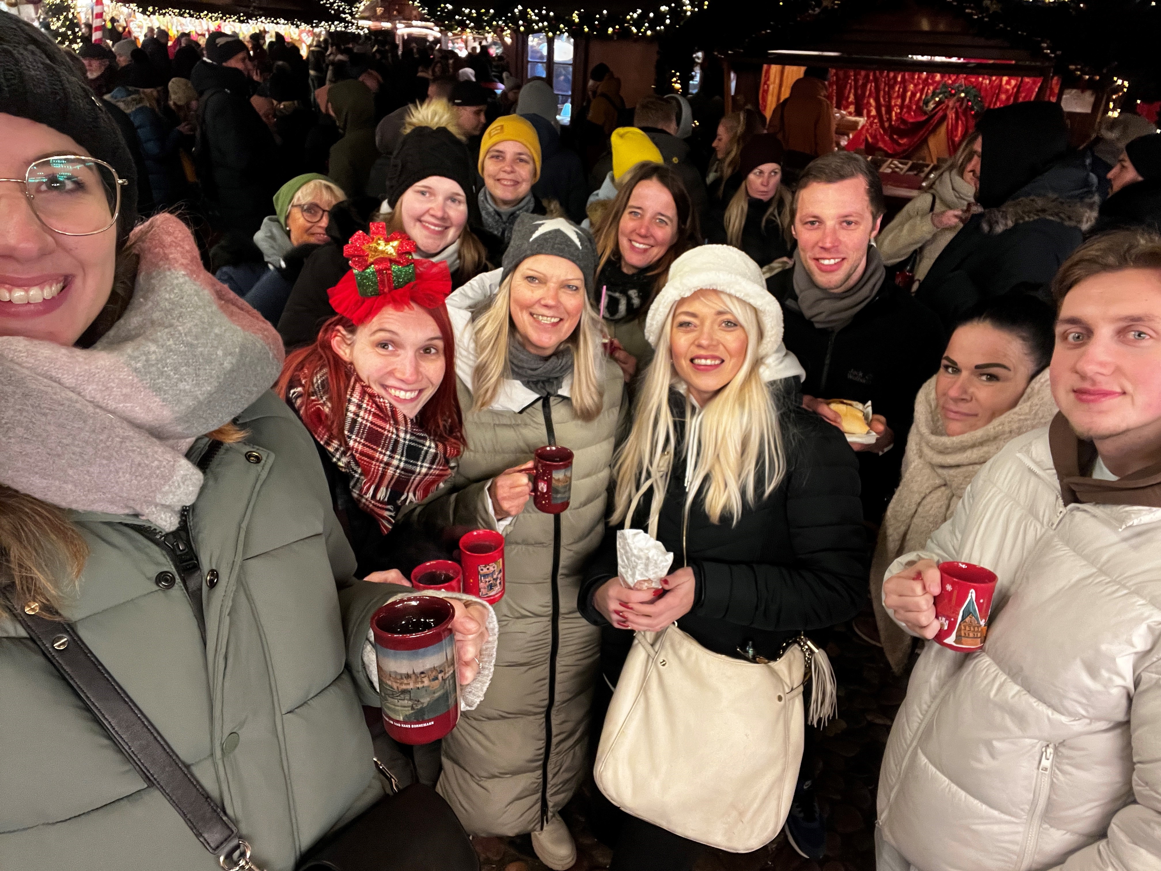 Eine größere Gruppe steht auf einem Weihnachtsmarkt. Eine junge Frau am linken Bildrand macht ein Selfie der Gruppe. 