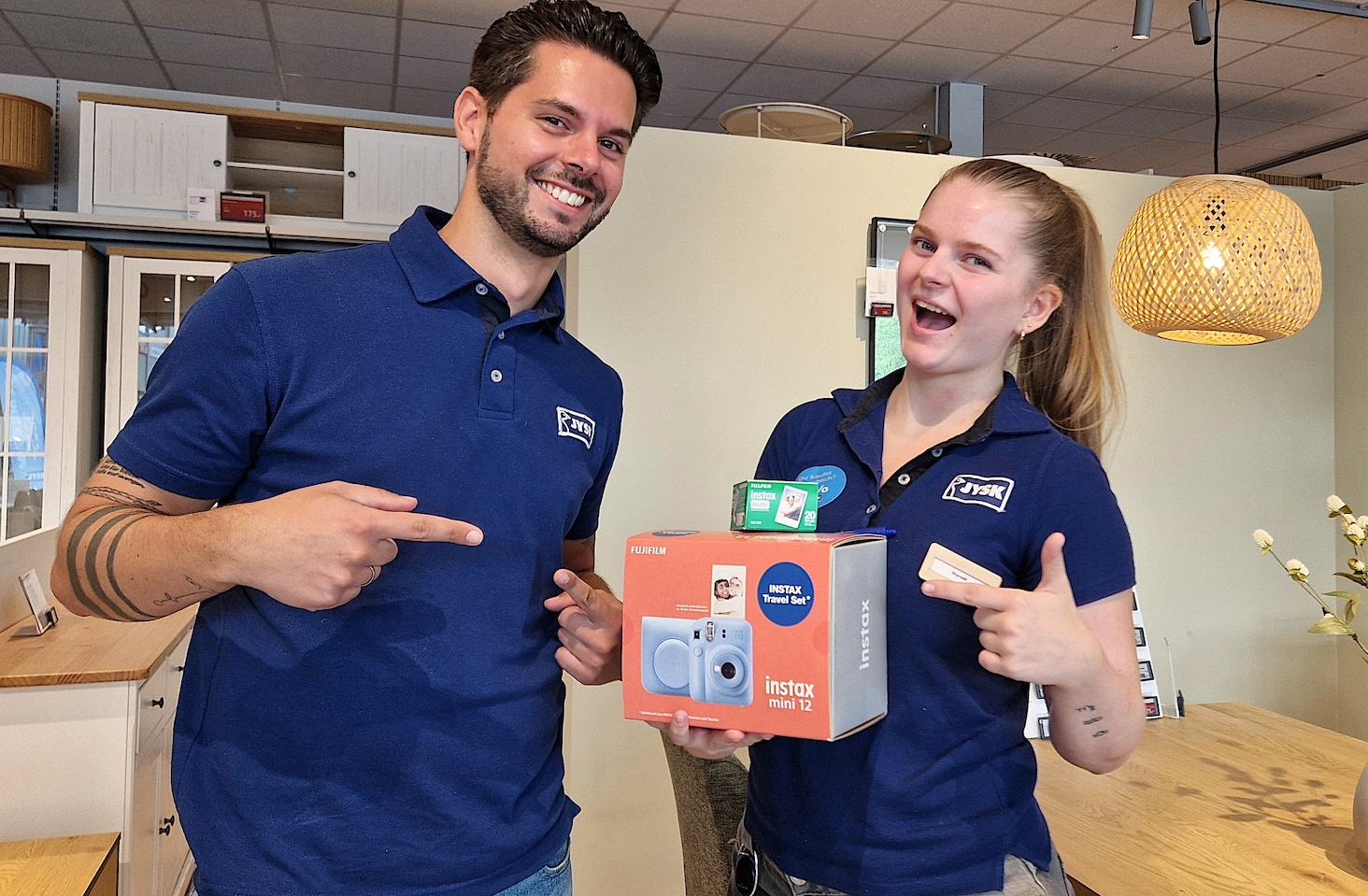 Ein Mann und eine Frau in Poloshirts stehen im Store. Die Frau hält einen Karton in der Hand. Der Mann und die Frau zeigen jeweils mit einem Finger auf diesen Karton. 