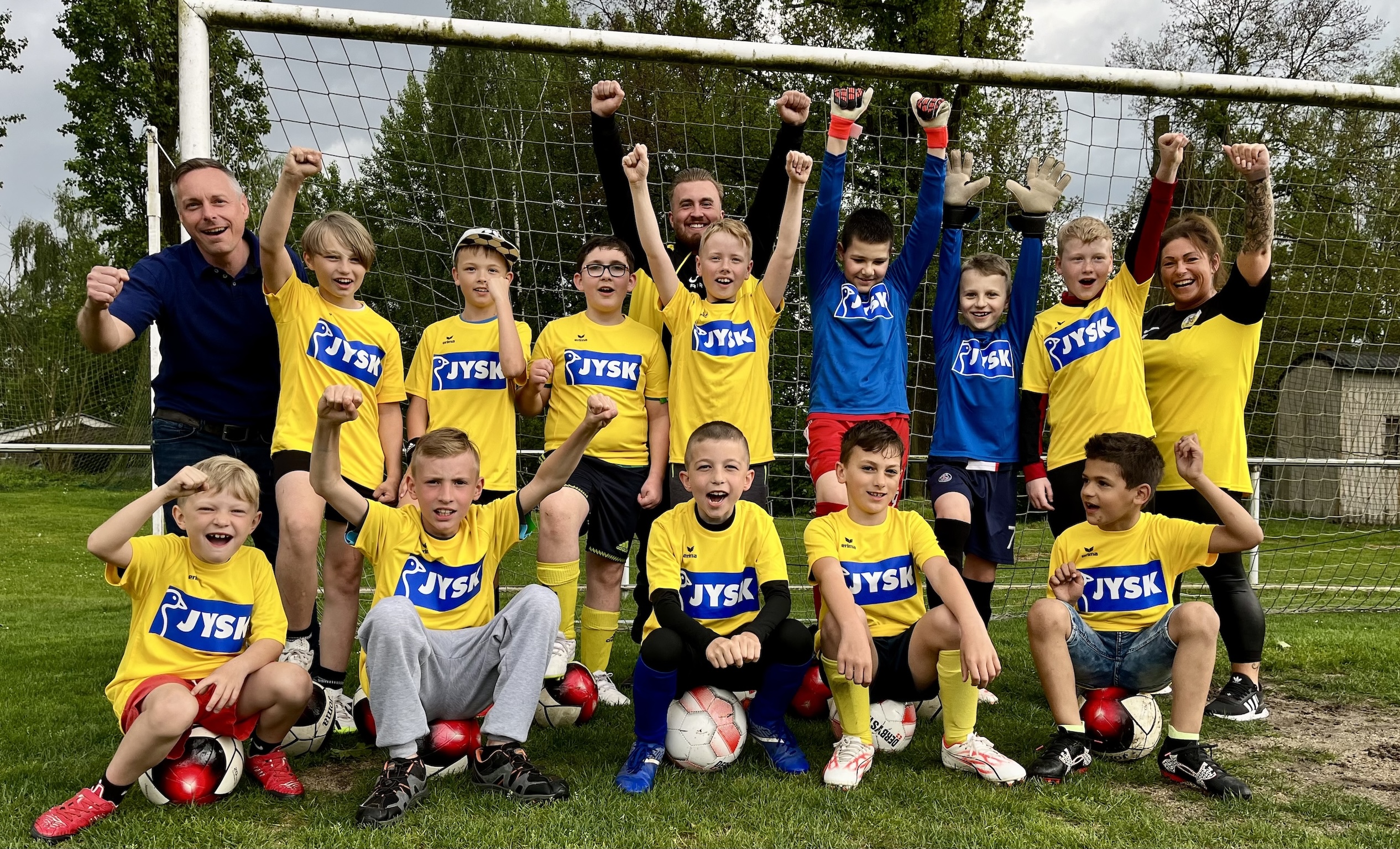 Viele Kinder sowie ein paar Erwachsene sitzen und stehen vor einem Fußballtor. Sie jubeln in die Kamera. 