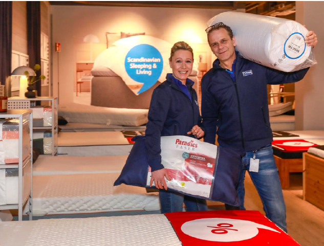 Ein Mann und eine Frau stehen in einem JYSK Store. Der Mann trägt eine Rollmatratze über die Schulter. De Frau hält ein Kissen in der Hand