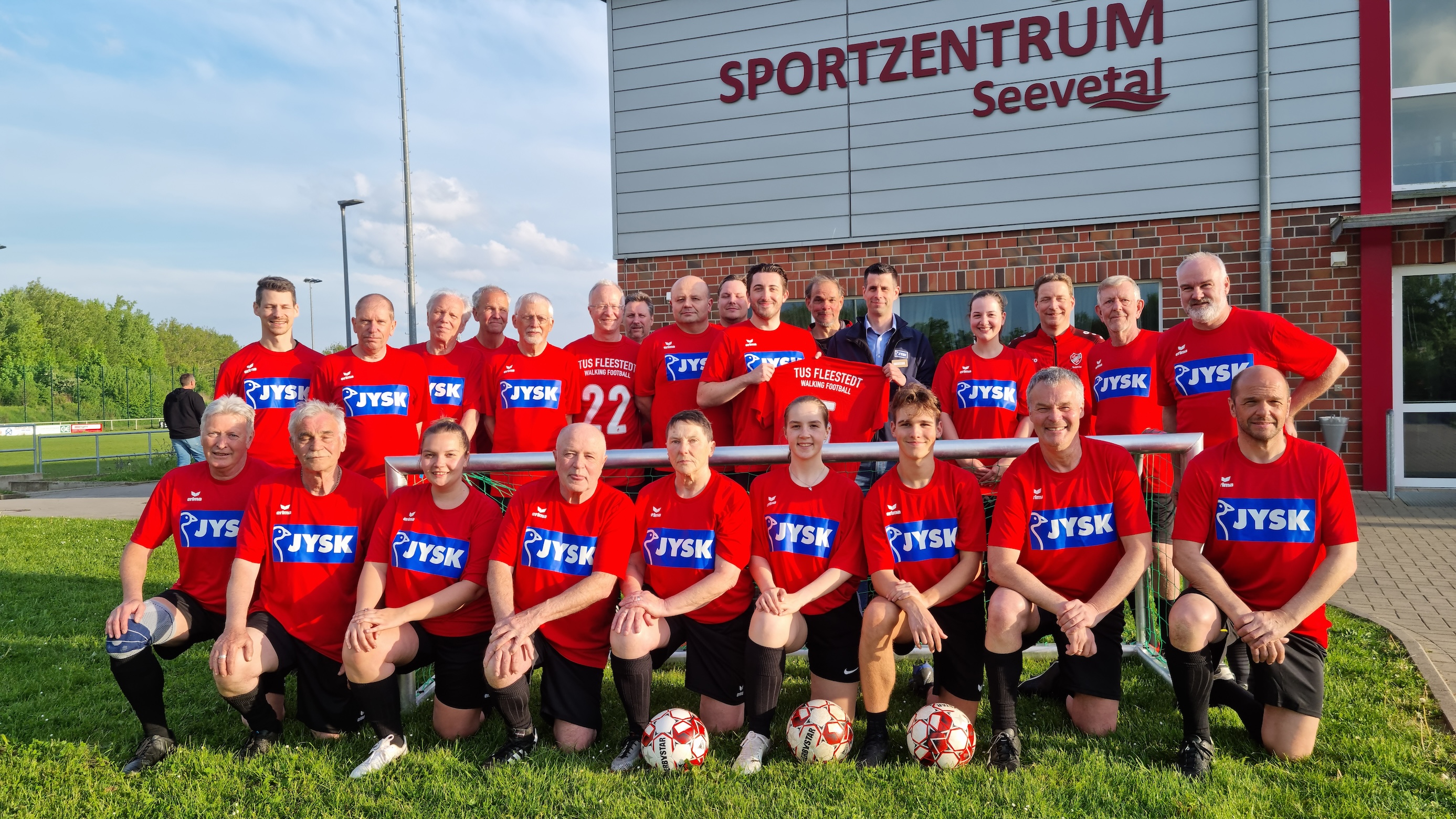 Viele Menschen – sowohl jung als auch alt – stehen und hocken auf einem Fußballplatz. Sie lächeln in die Kamera. Im Hintergrund ist ein Clubhaus zu erkennen.