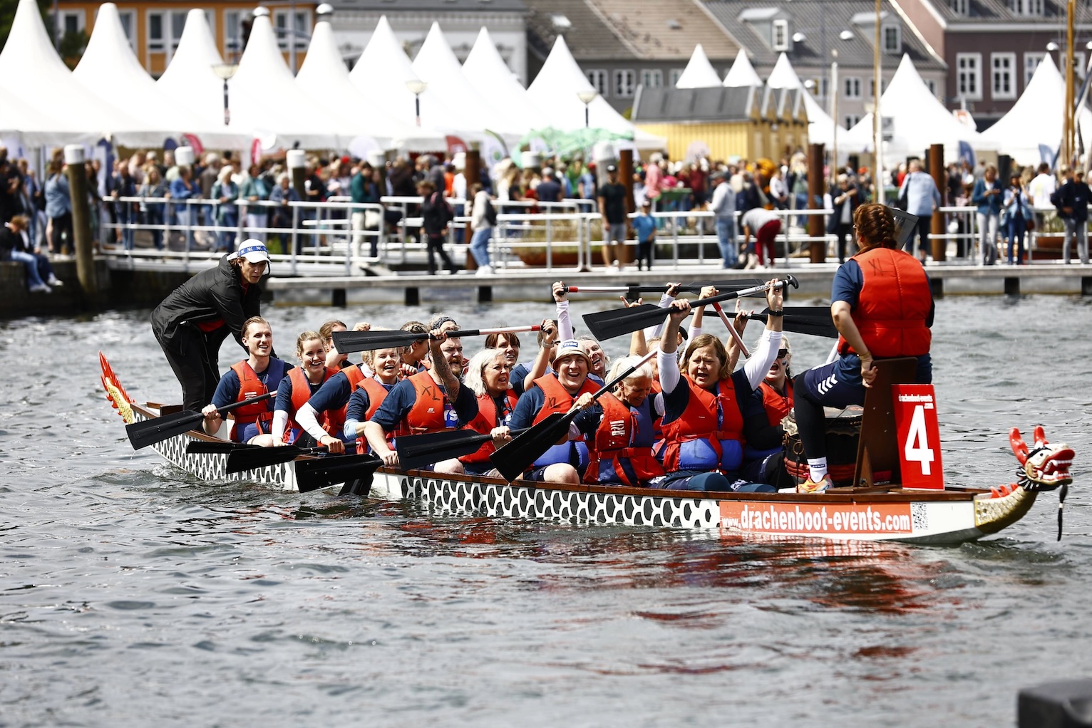 19 Menschen sitzen in einem Ruderboot, eine weitere Person steht auf dem Boot. Viele der Menschen lachen und strecken ihre Ruder in die Höhe
