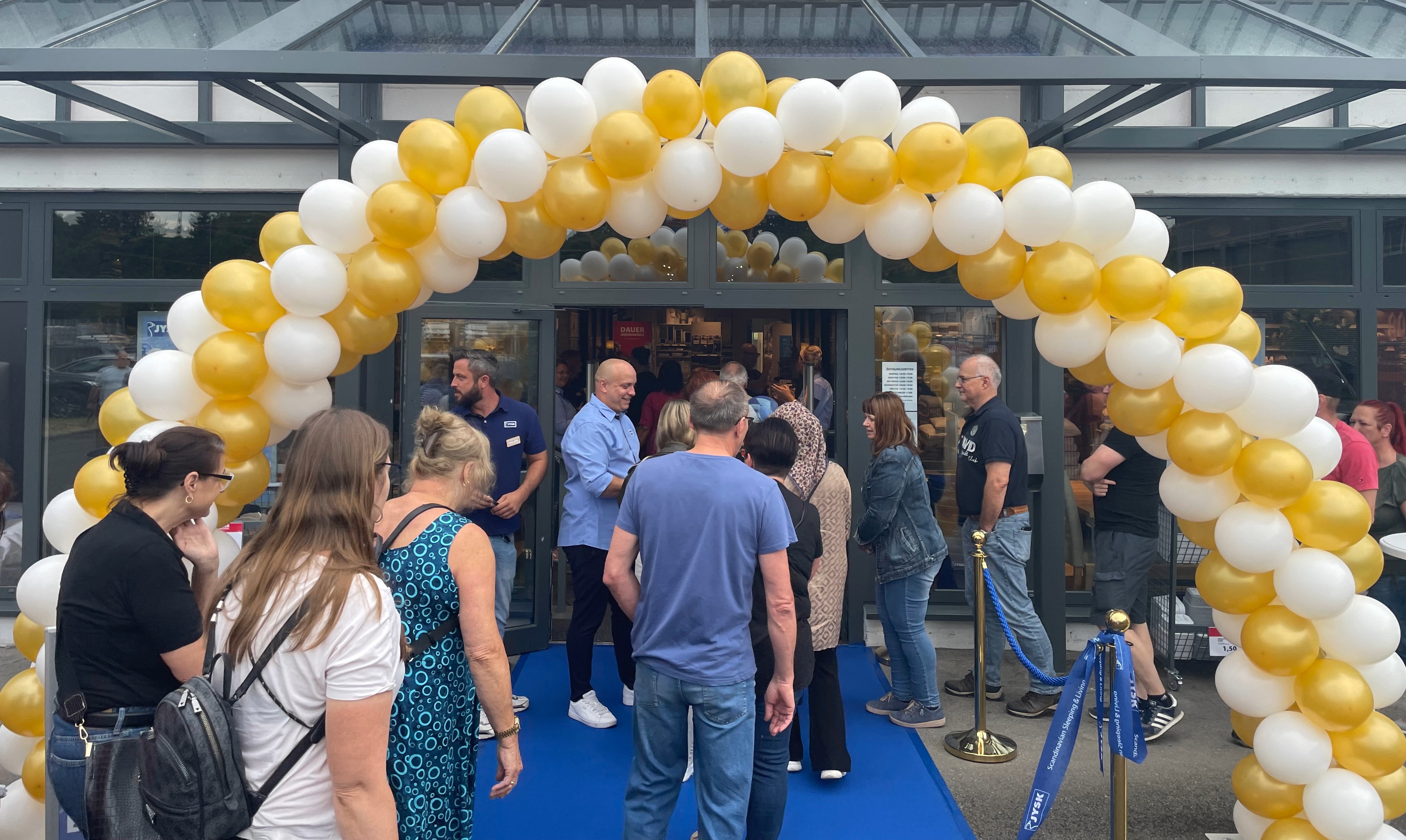 Sehr viele Menschen stehen vor einem JYSK Store oder gehen gerade rein. Eine Luftballongirlande schmückt den Eingang. 