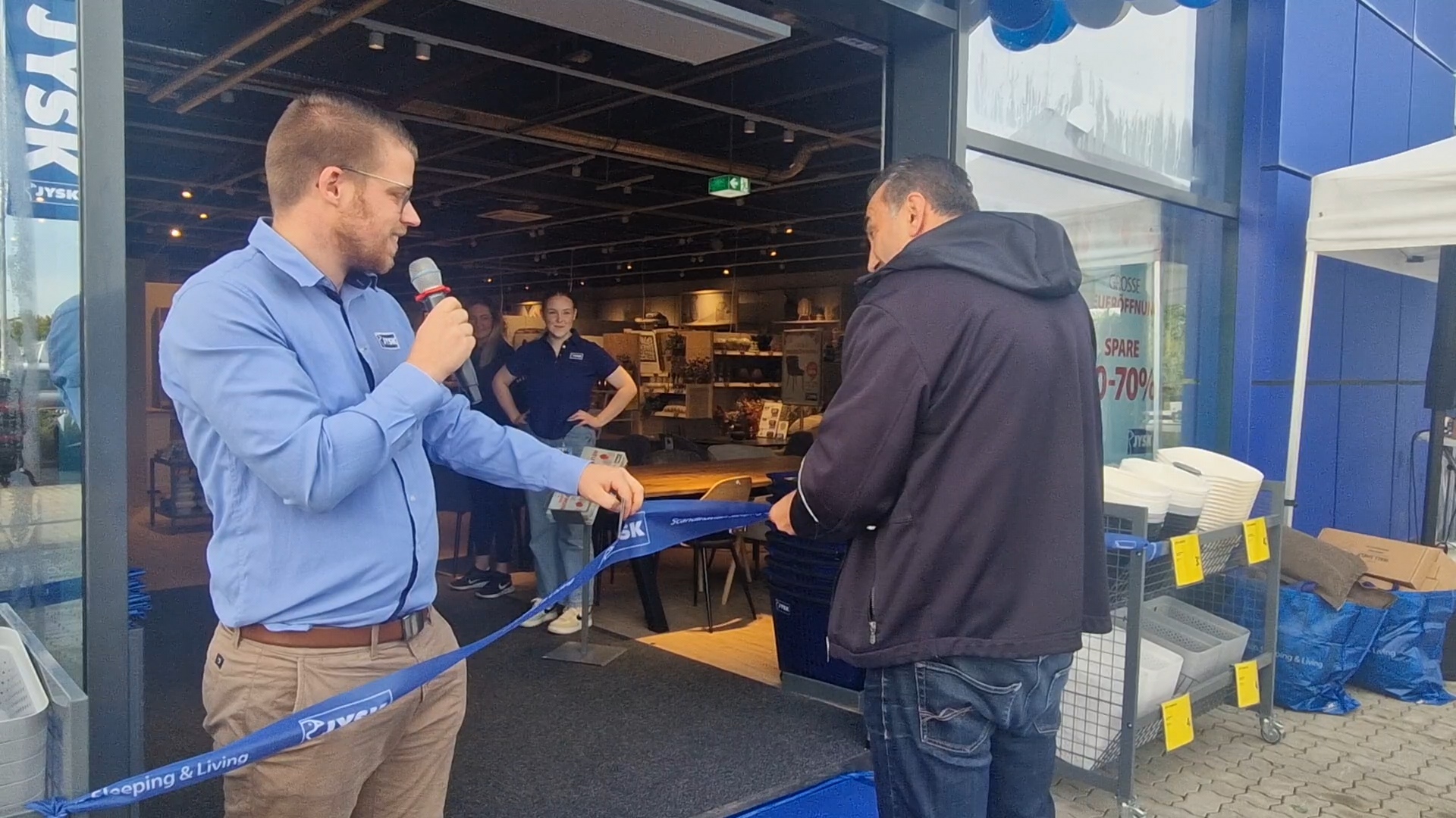 Zwei Männer stehen vor einem JYSK Store. Einer hält in der einen Hand ein Mikrofon und in der anderen ein dickeres Band. Der zweite Mann hält eine Schere in der Hand. 