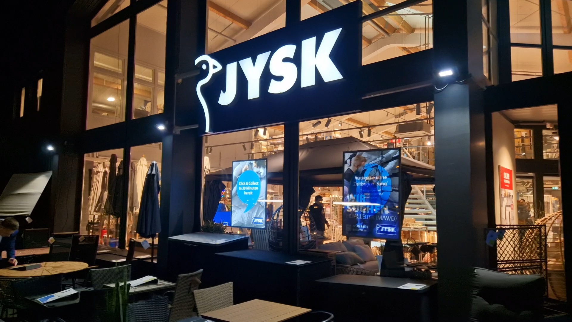 Ein JYSK-Store mit großer Fensterfront von außen. Es ist bereits dunkel und das JYSK-Schild leuchtet.