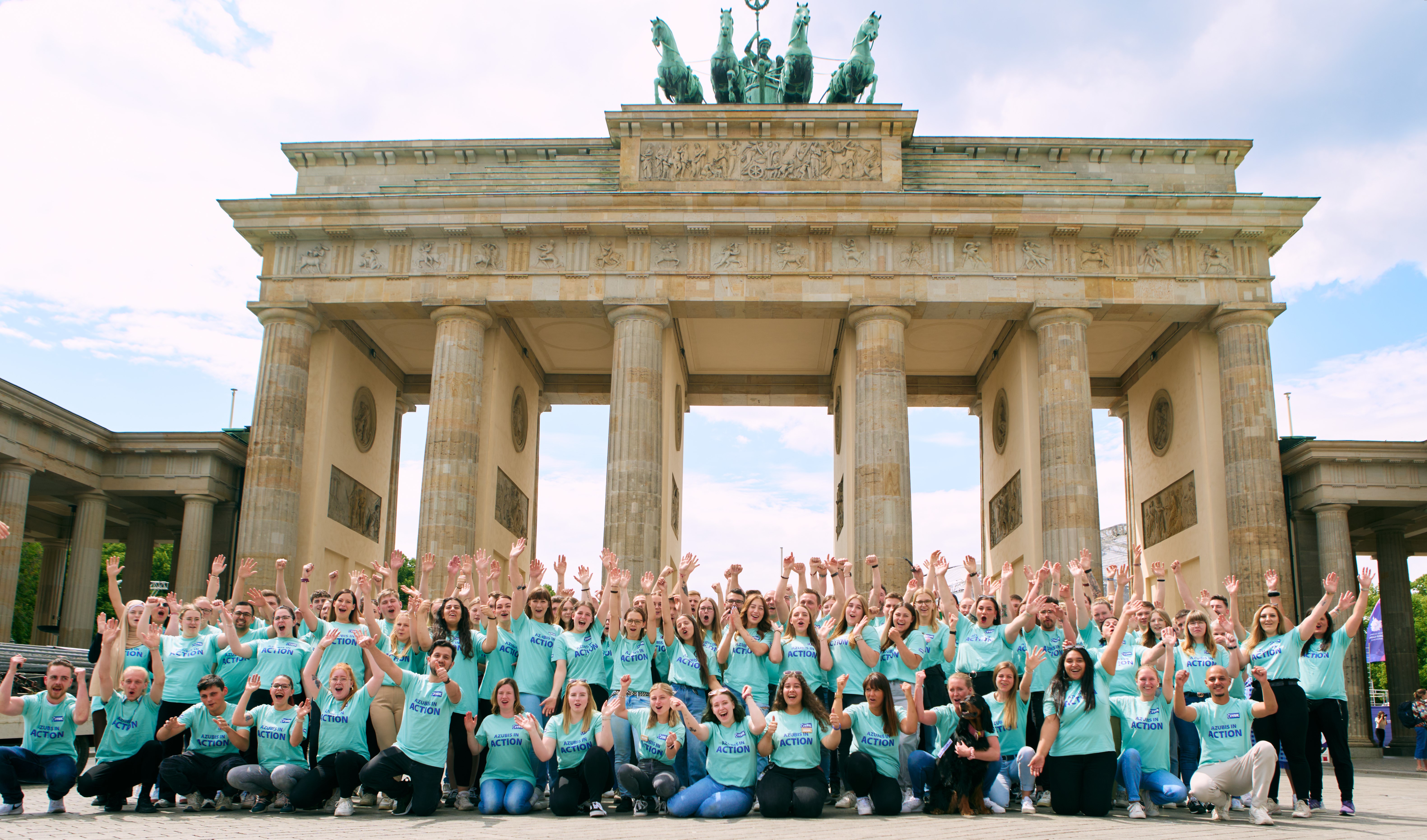 Mehr als 100 junge Menschen stehen vorm Brandenburger Tor – ein Tor, das auf sechs Säulen steht und sich mitten in Berlin befindet. Die Menschen haben die Arme hochgerissen und jubeln.