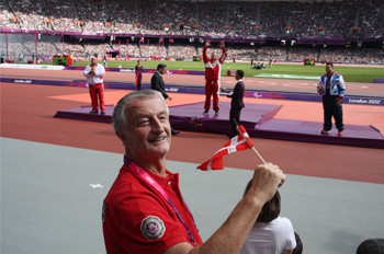 Ein Mann lacht in die Kamera. Er hält ein dänisches Fähnchen in der Hand. Er sitzt in einem Stadion. im Hintergrund ist eine Siegerehrung zu erkennen. 