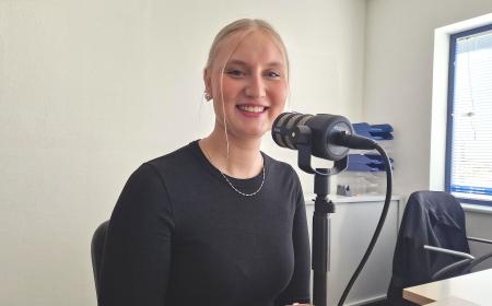 Eine junge Frau sitzt vor einem Mikrofon. Ihre langen Haare sind zu einem Zopf gebunden. Sie trägt eine Kette um den Hals und ein langärmeliges Oberteil. Die Frau lächelt in die Kamera.