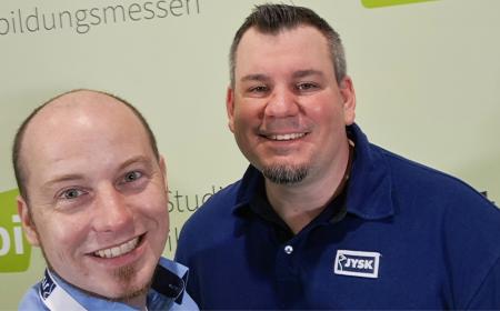 Zwei Männer in JYSK-Kleidung machen ein Selfie und lachen in die Kamera
