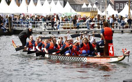 19 Menschen sitzen in einem Ruderboot, eine weitere Person steht auf dem Boot. Viele der Menschen lachen und strecken ihre Ruder in die Höhe