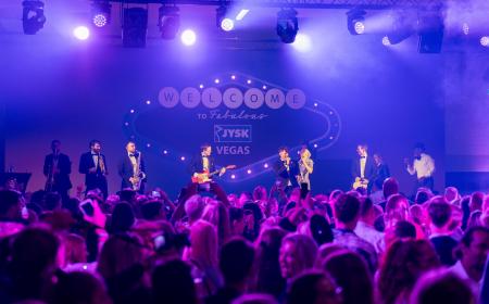 Eine Band steht auf einer Bühne und performt. Davor tanzen und stehen zahlreiche Menschen